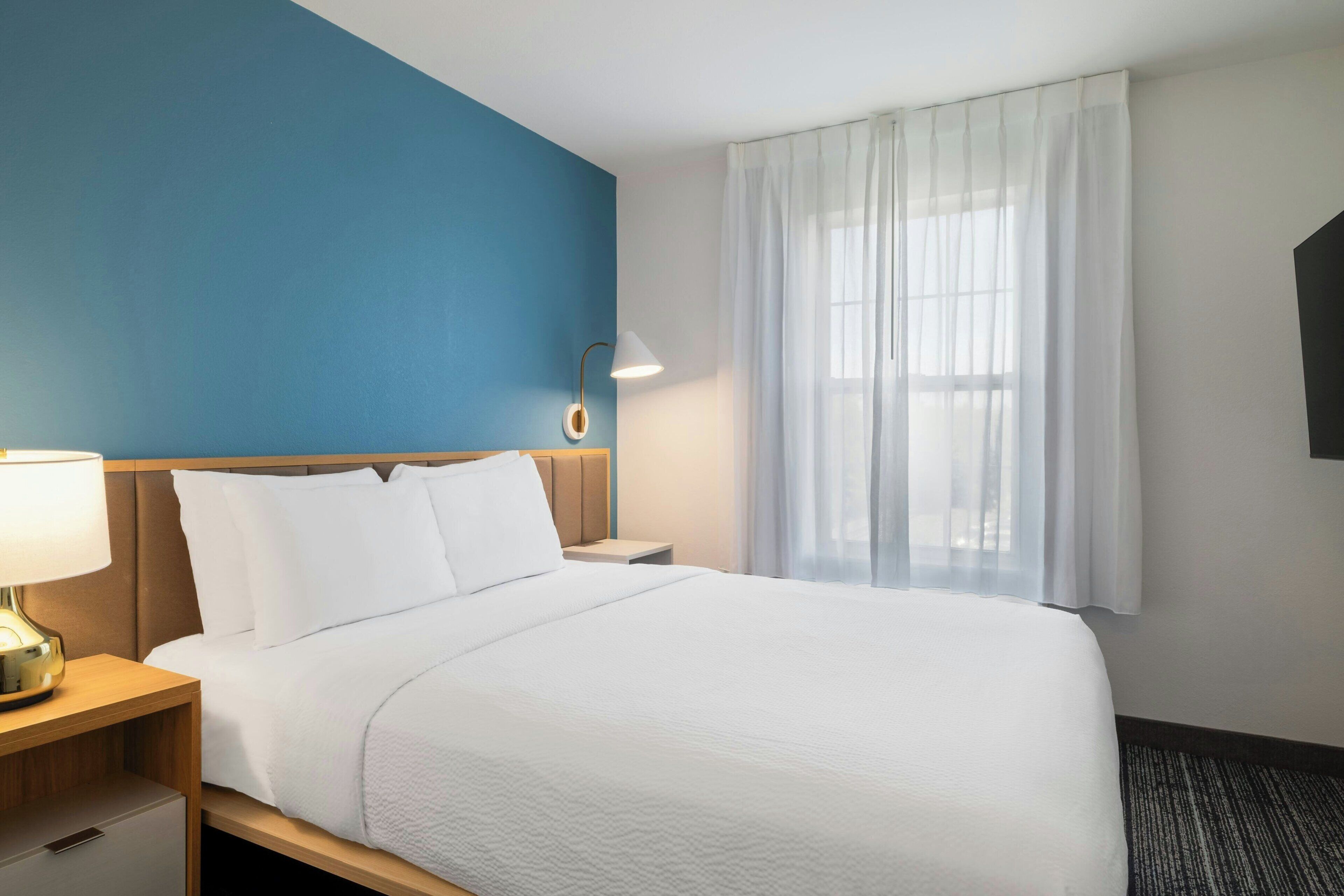 Una habitación de hotel con una cama, una mesita de noche, una lámpara, un televisor y una ventana con cortinas.