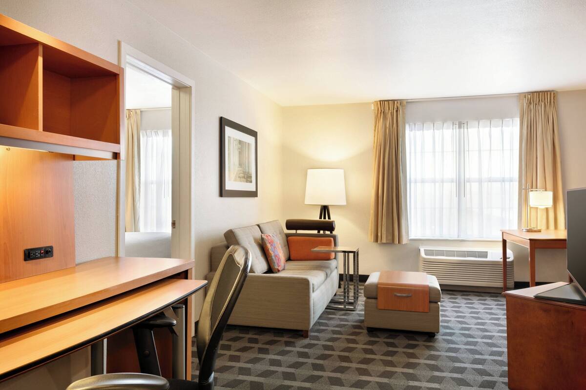 Et hotelværelse med sofa, skrivebord, stol, lampe, fjernsyn og et vindue med gardiner.