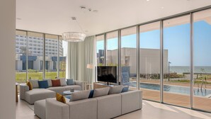 A modern living room with a large sofa, a flat-screen TV, and a view of a pool and ocean. - Dusit Ajman Resort & Villas (Ajman)