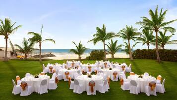 A beachfront wedding setup with round tables and chairs, arranged on a green lawn with palm trees and the ocean in the background.