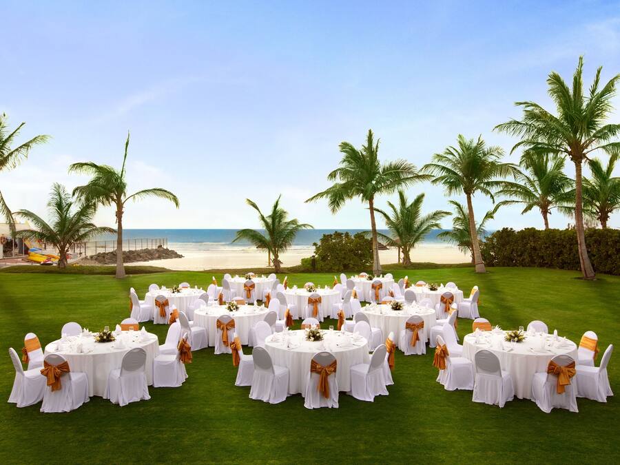 A beachfront wedding setup with round tables and chairs, arranged on a green lawn with palm trees and the ocean in the background.
