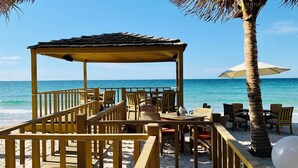 A beachside restaurant with outdoor seating, a thatched roof structure, and a view of the ocean. - Dusit Ajman Resort & Villas (Ajman)