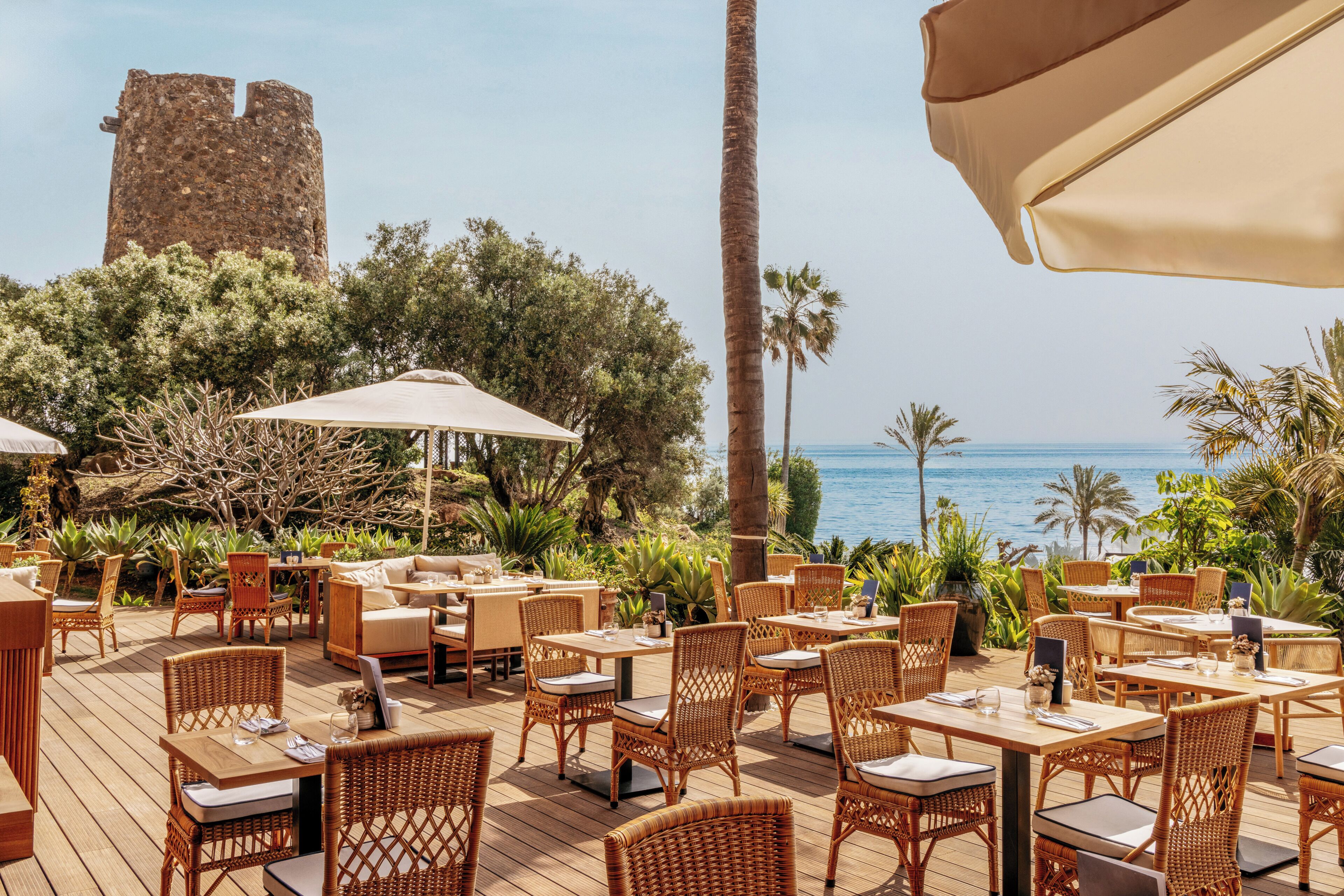 An outdoor seating area with wicker furniture and umbrellas, overlooking a body of water with a historic tower in the background.