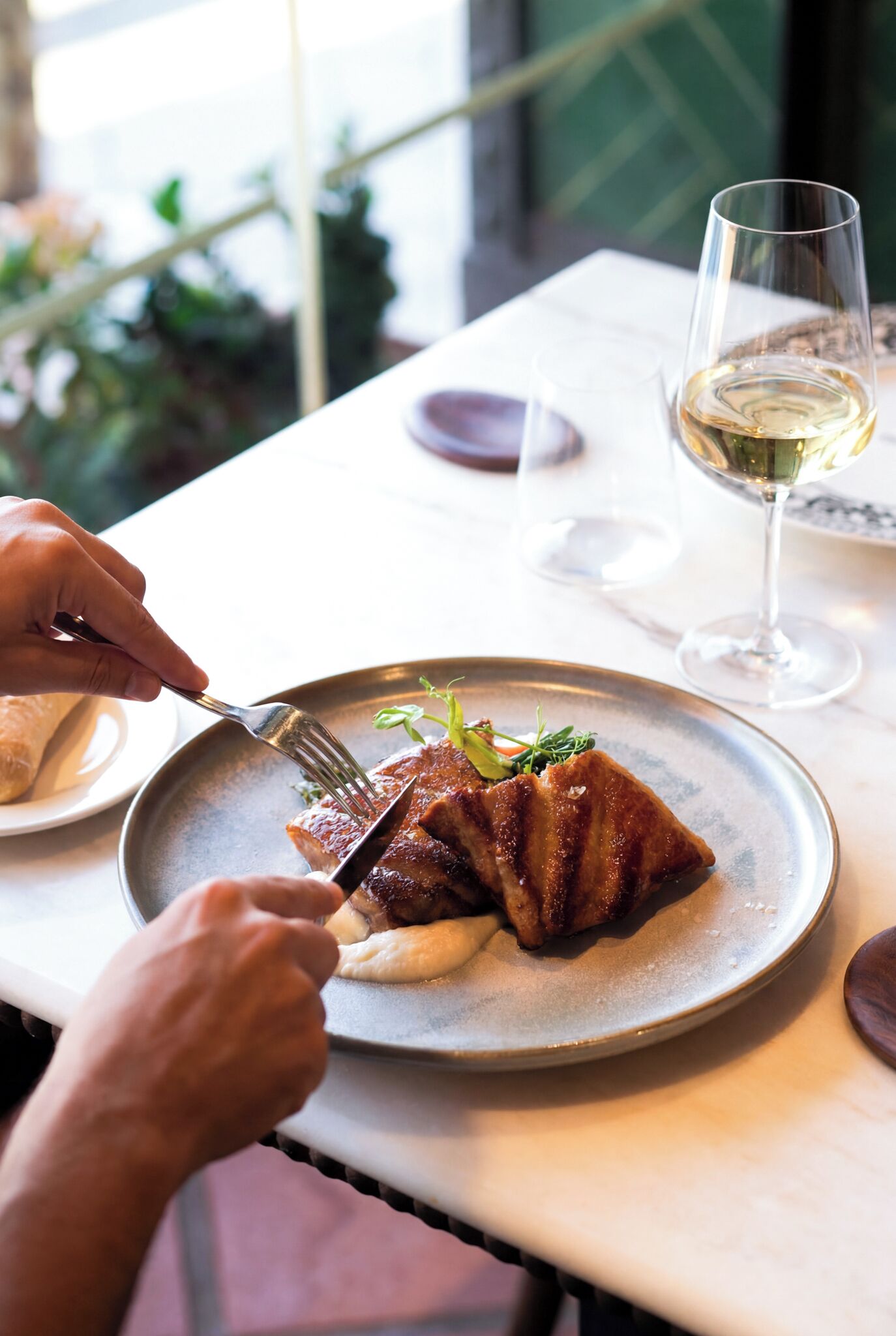 Una persona cenando en una mesa con un plato de comida, una copa de vino blanco y un tenedor.