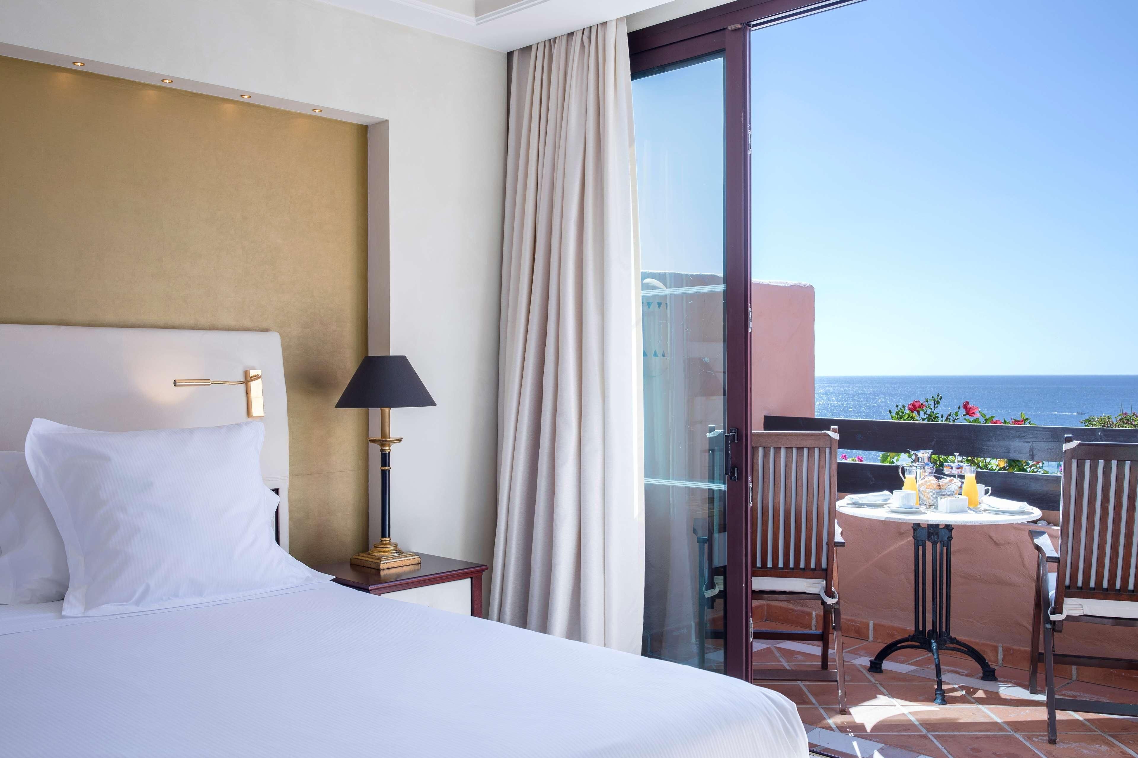 A hotel room with a bed, a nightstand, a lamp, a table with chairs, and a view of the sea.