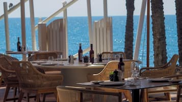 Un espacio de comedor al aire libre con mobiliario de mimbre, vistas al mar y un entorno tropical con palmeras.