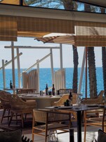 Un espacio de comedor al aire libre con mobiliario de mimbre, vistas al mar y un entorno tropical con palmeras.