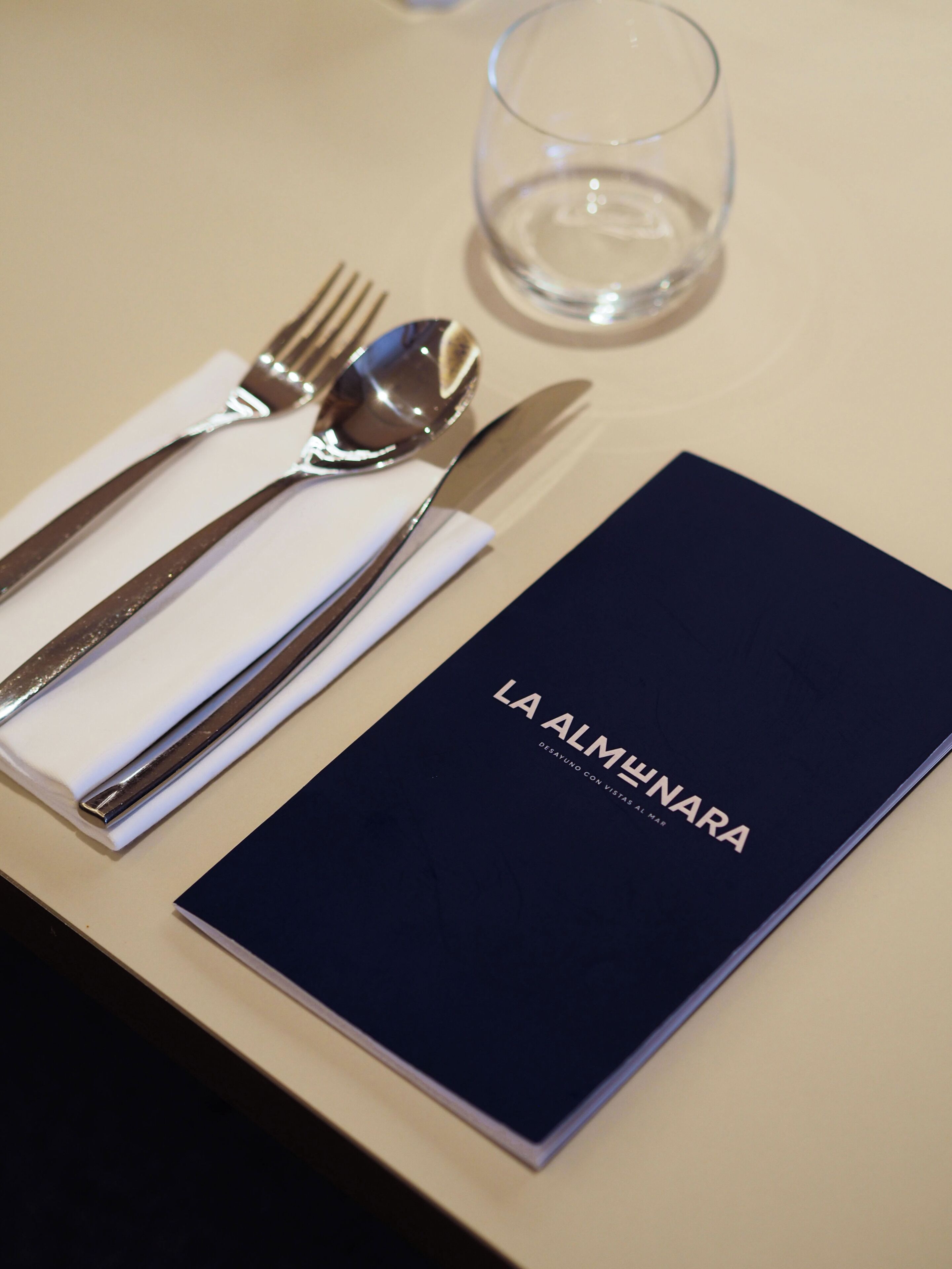 A table setting with a folded napkin, cutlery, and a glass of water. A menu titled 'LA ALMENARA' is also visible.