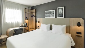 Une chambre d’hôtel avec un grand lit, un bureau avec une chaise, un porte-manteau et deux lampes fixées au mur.