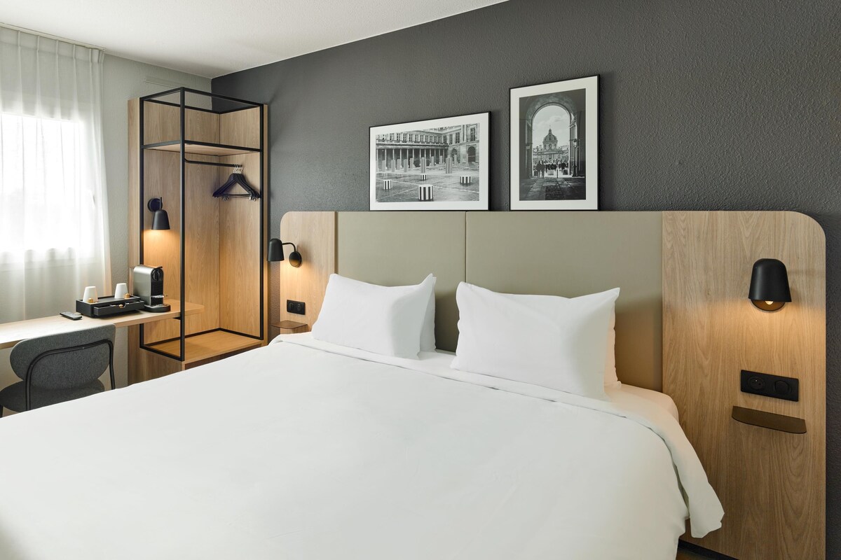 Une chambre d’hôtel avec un grand lit, un bureau avec une chaise, un porte-manteau et deux lampes fixées au mur.