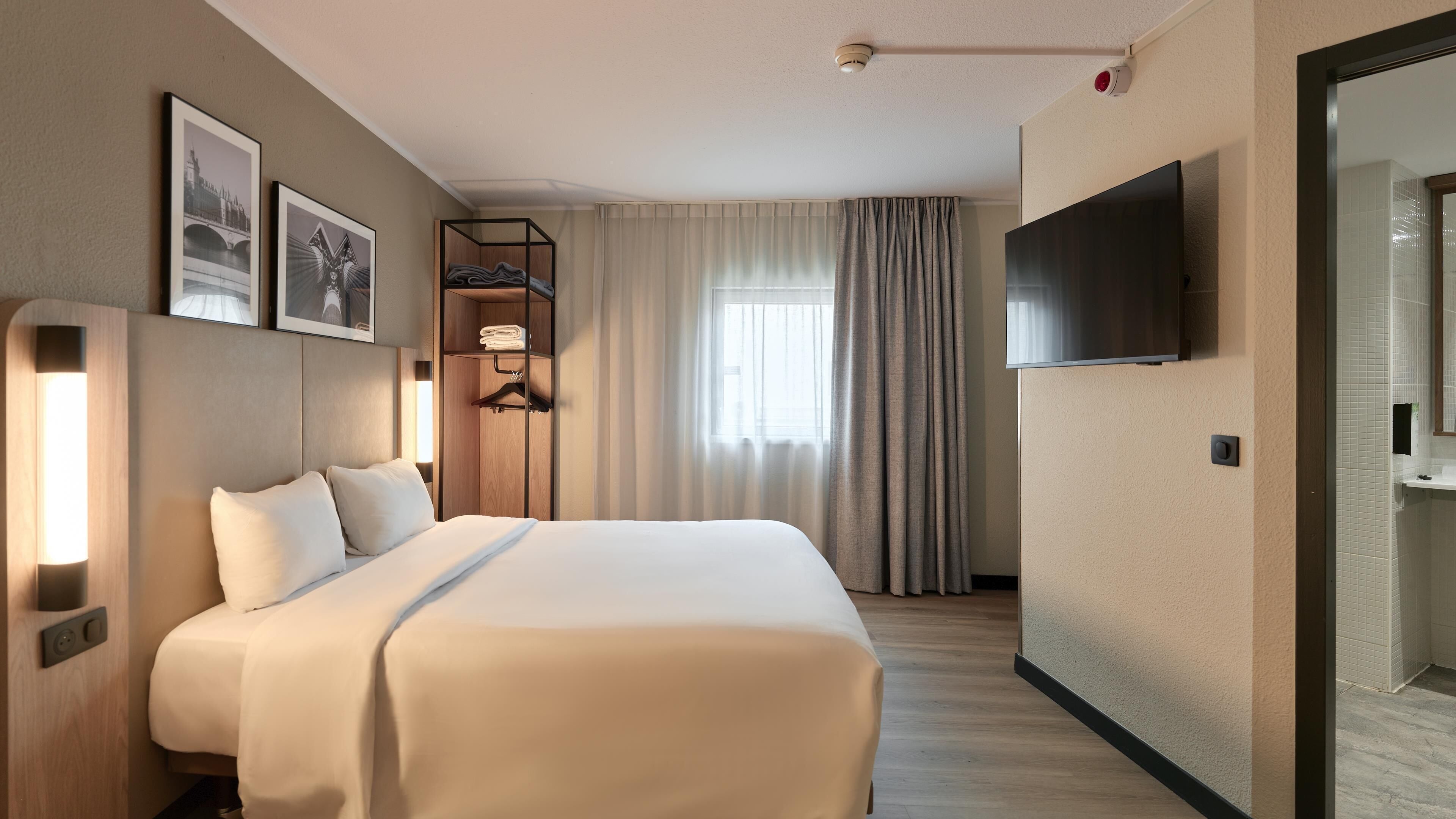 A hotel room with a bed, a TV, a wardrobe, a mirror, and a window with curtains.