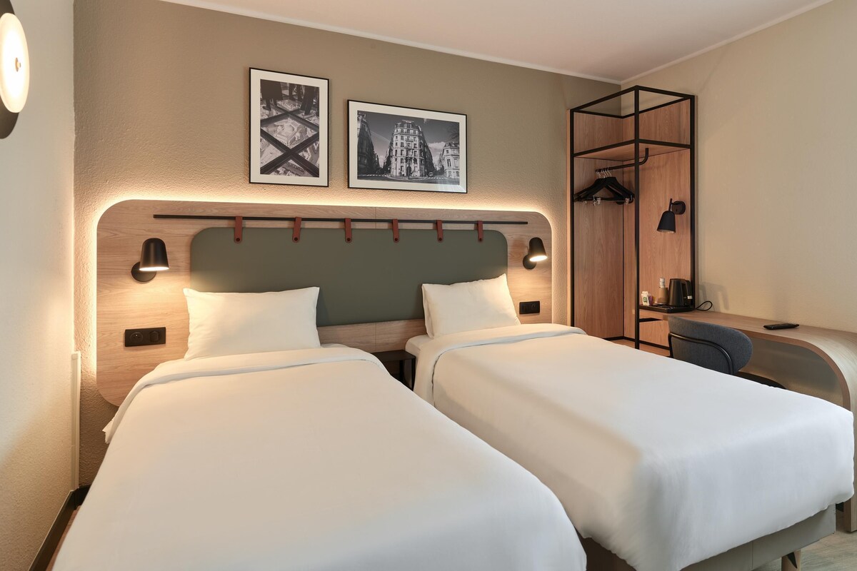Ett hotellrum med två sängar, ett skrivbord med en stol, en garderob och två väggmonterade lampor.