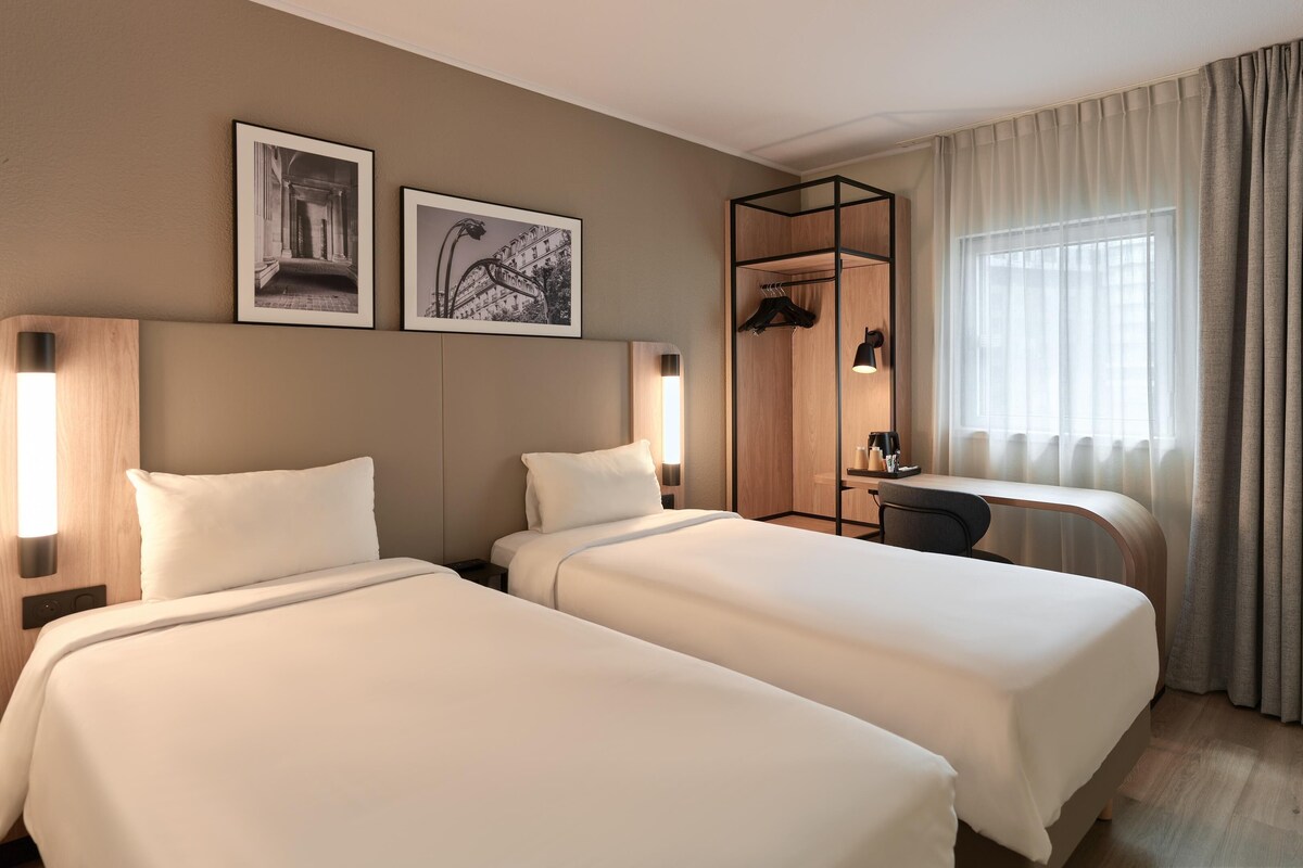 Ett hotellrum med två sängar, ett skrivbord, en stol, en garderob och två inramade tavlor på väggen.