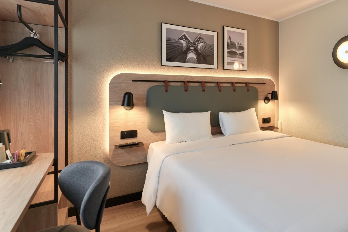 Une chambre d’hôtel avec un lit, des tables de chevet, un bureau, une chaise et deux tableaux encadrés au mur.