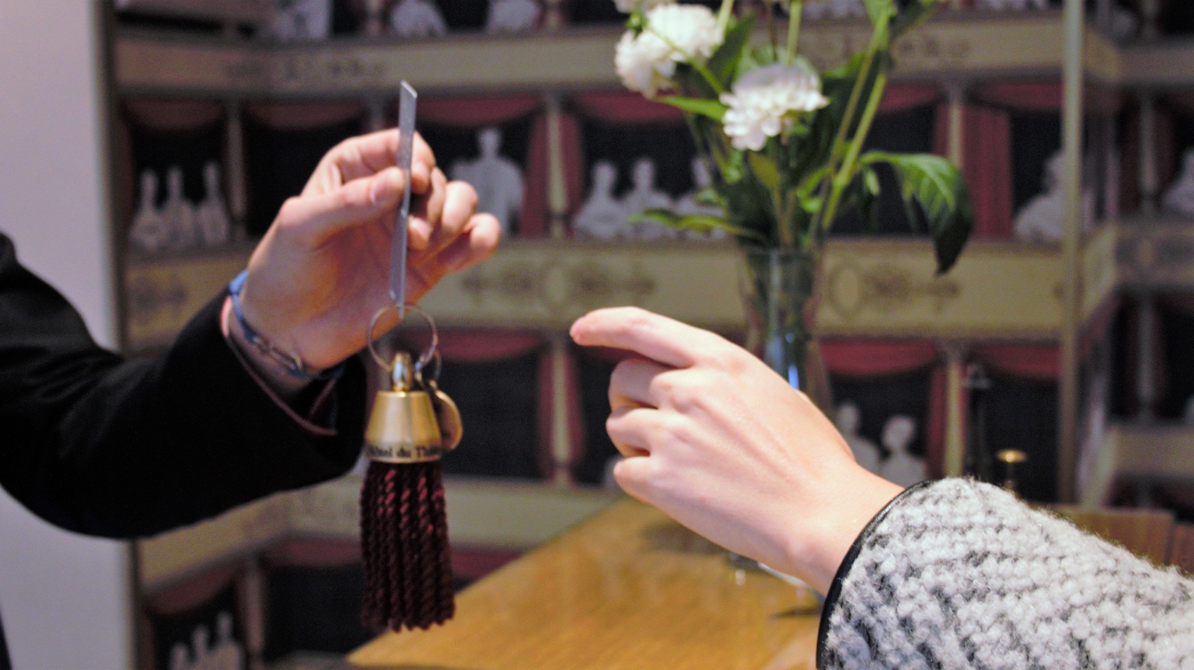A person holding a key and a bottle with a tassel, with another hand pointing at them.