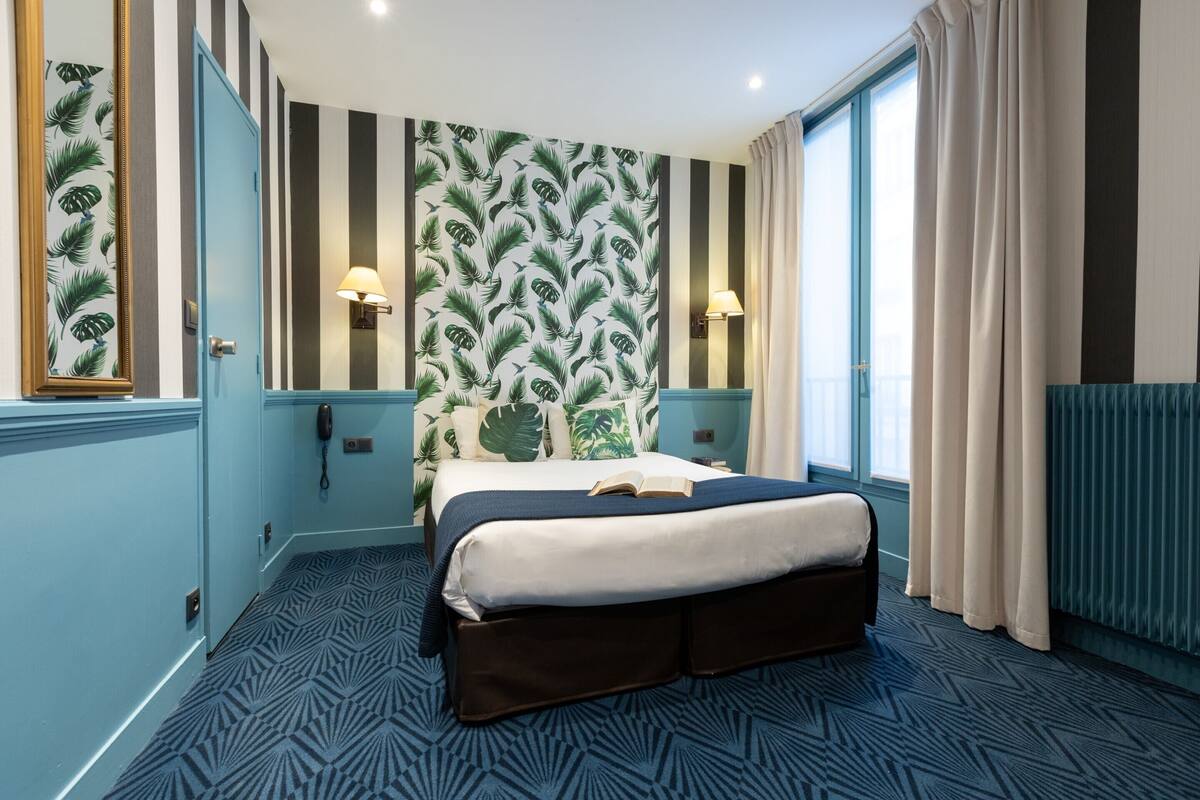 Une chambre d’hôtel avec un lit, une grande fenêtre avec des rideaux, un mur orné d’un motif de feuilles et une moquette à motifs.