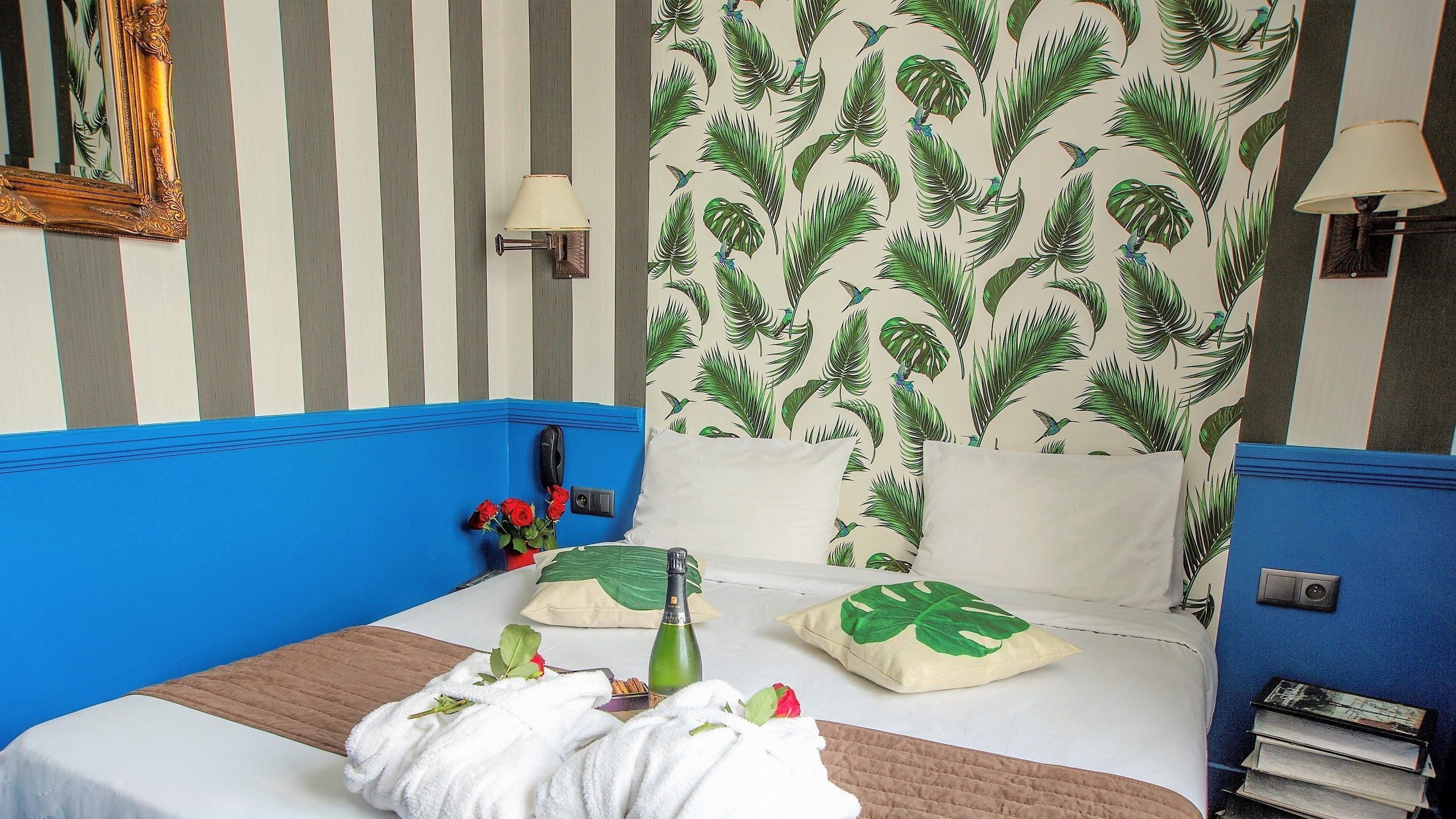 A hotel room with a striped wall, a bed with a blue headboard, a floral-patterned curtain, and a bedside table with a bottle and flowers.