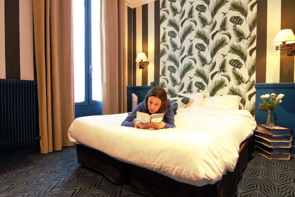 Une chambre d’hôtel avec un lit, une table de chevet ornée d’un vase de fleurs, une lampe et un mur décoré d’un motif de feuilles tropicales.
