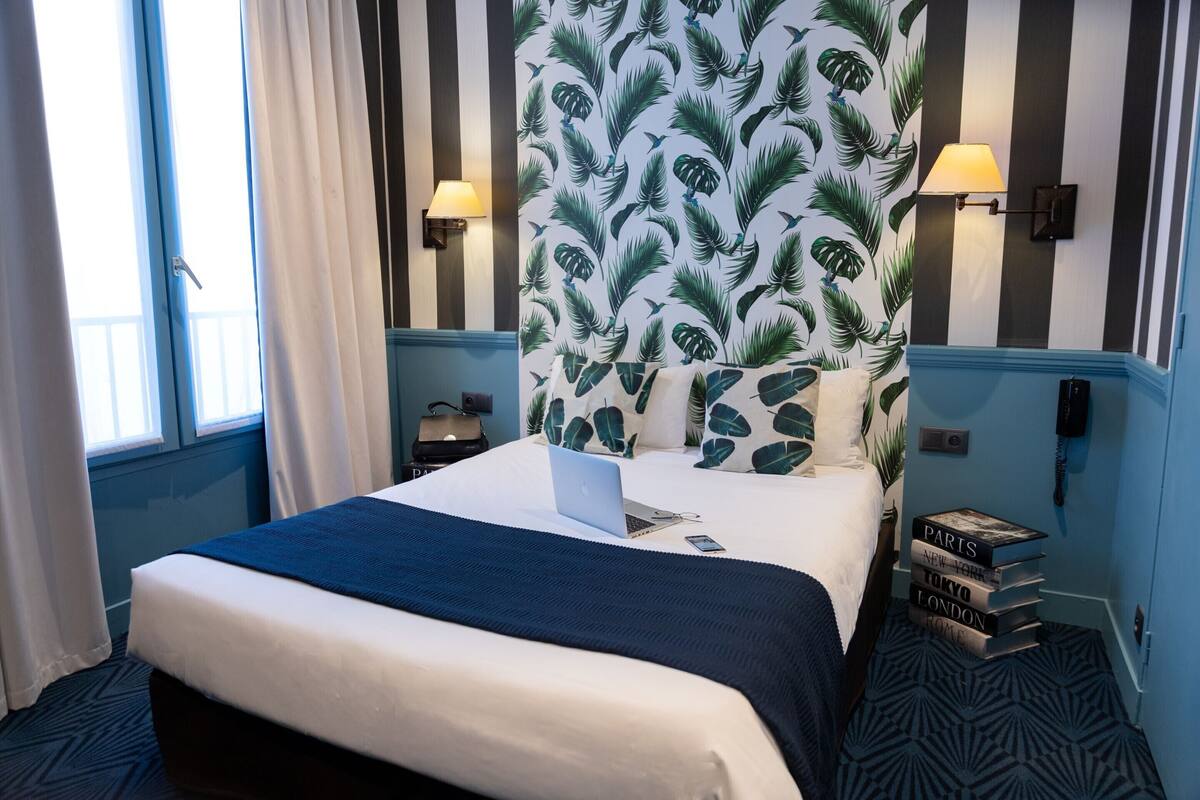 Une chambre d’hôtel avec un lit, un ordinateur portable, une pile de livres et un mur orné d’un motif de feuilles.