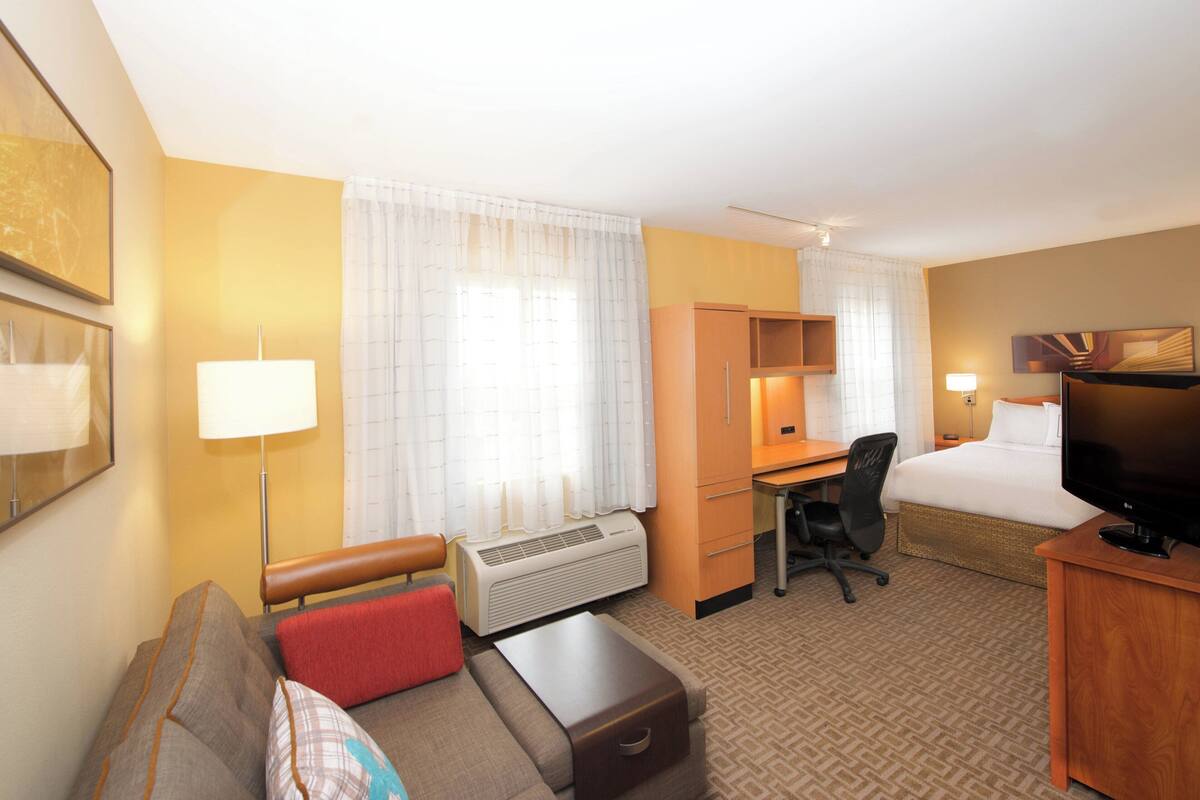 Habitación de hotel con cama, escritorio, silla, sofá, televisión, lámpara y ventana con cortinas.