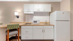 Una cocina compacta con armarios blancos, microondas, refrigerador, lavamanos y una silla con cojín verde.