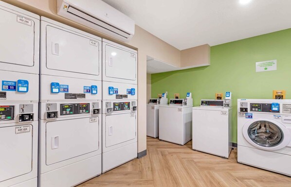 A laundry facility with multiple washing machines and dryers, a green wall, and a sign indicating laundry products can be purchased at the front desk.