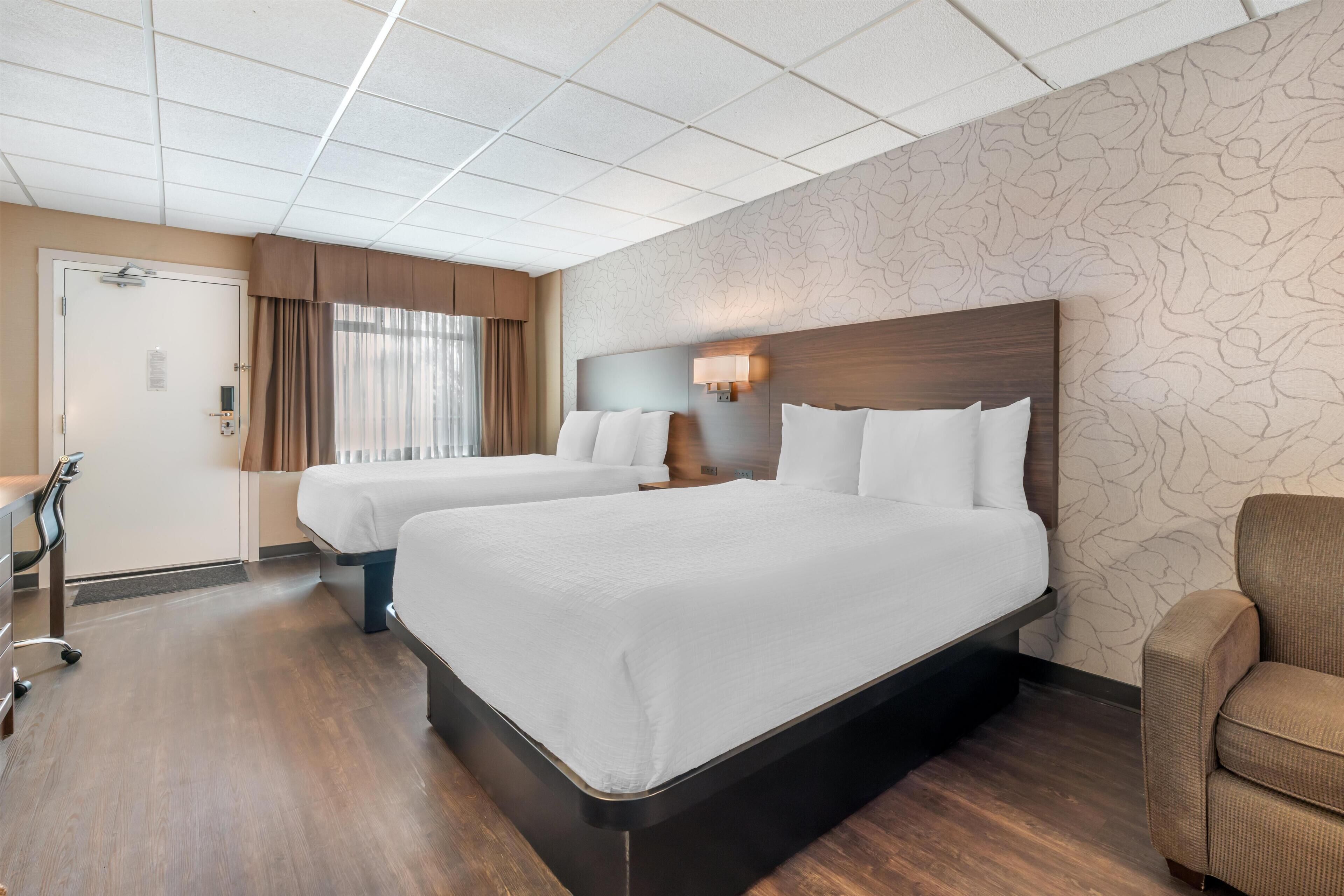 A hotel room with two beds, a desk, a chair, a window with curtains, and a patterned wall.