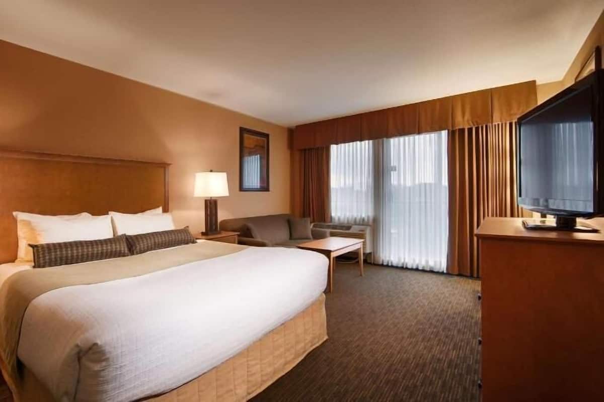 Una habitación de hotel con una cama grande, dos mesitas de noche con lámparas, un escritorio con silla, un televisor y un ventanal con cortinas.