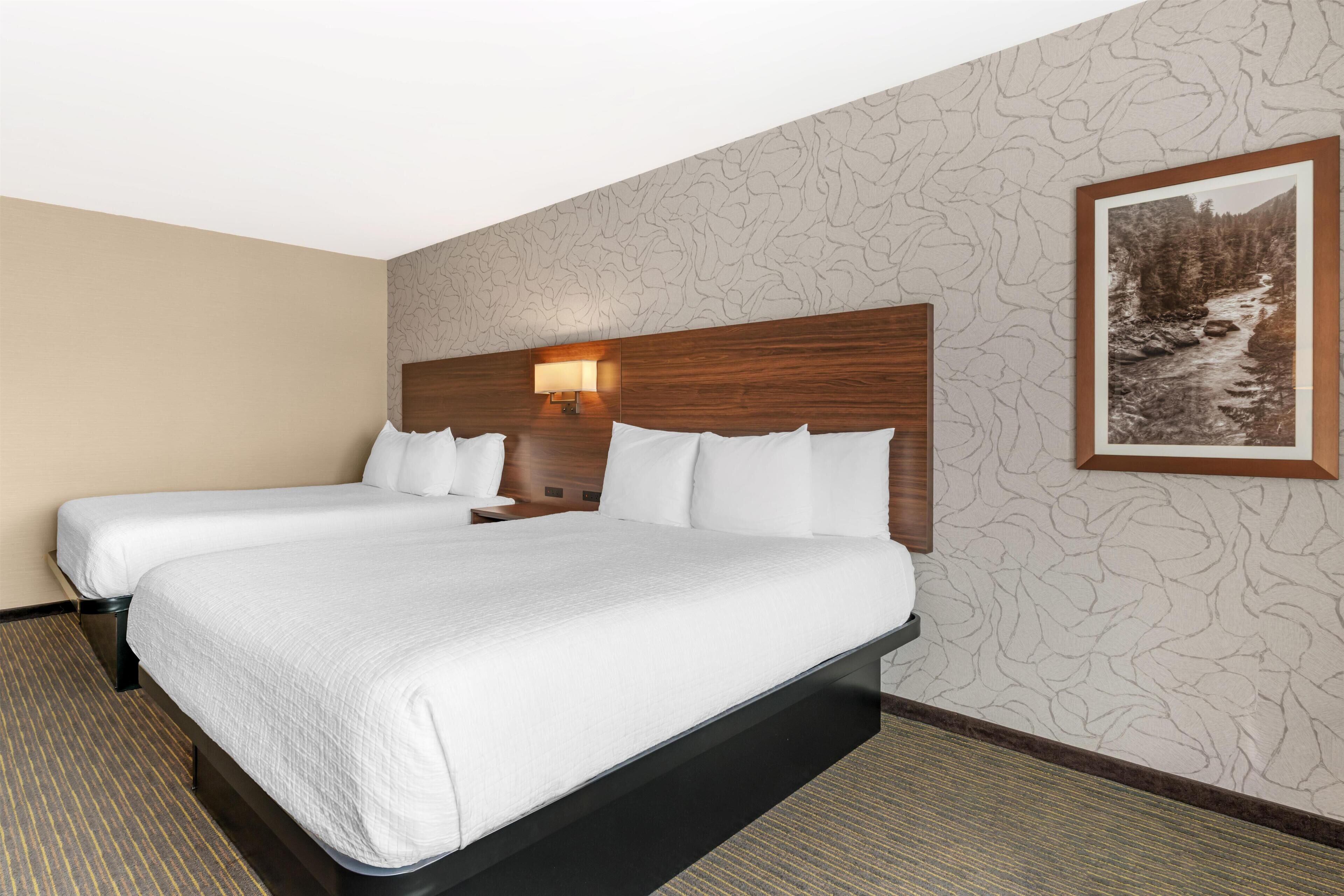 Habitación de hotel con dos camas, cabecero de madera, lámpara de pared e imagen enmarco de un paisaje.