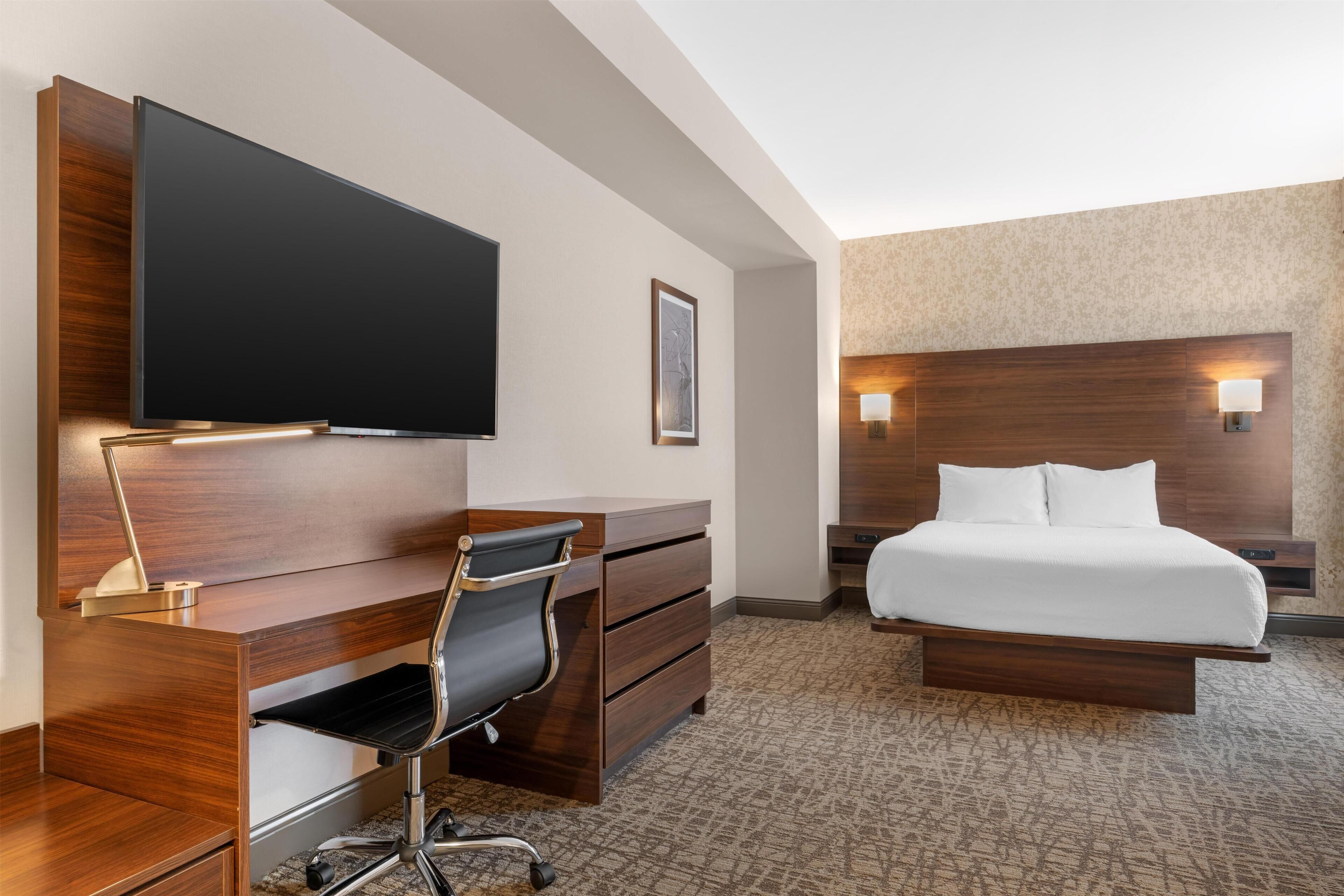 A hotel room with a wooden desk, a flat-screen TV, a bed with white linens, and a patterned carpet.