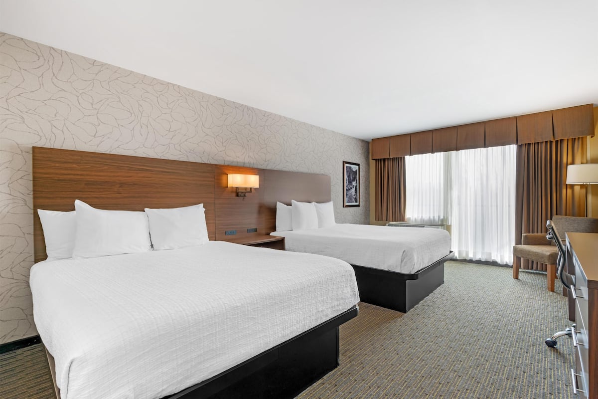 Habitación de hotel con dos camas, un escritorio, una silla, una lámpara y un ventanal con cortinas.