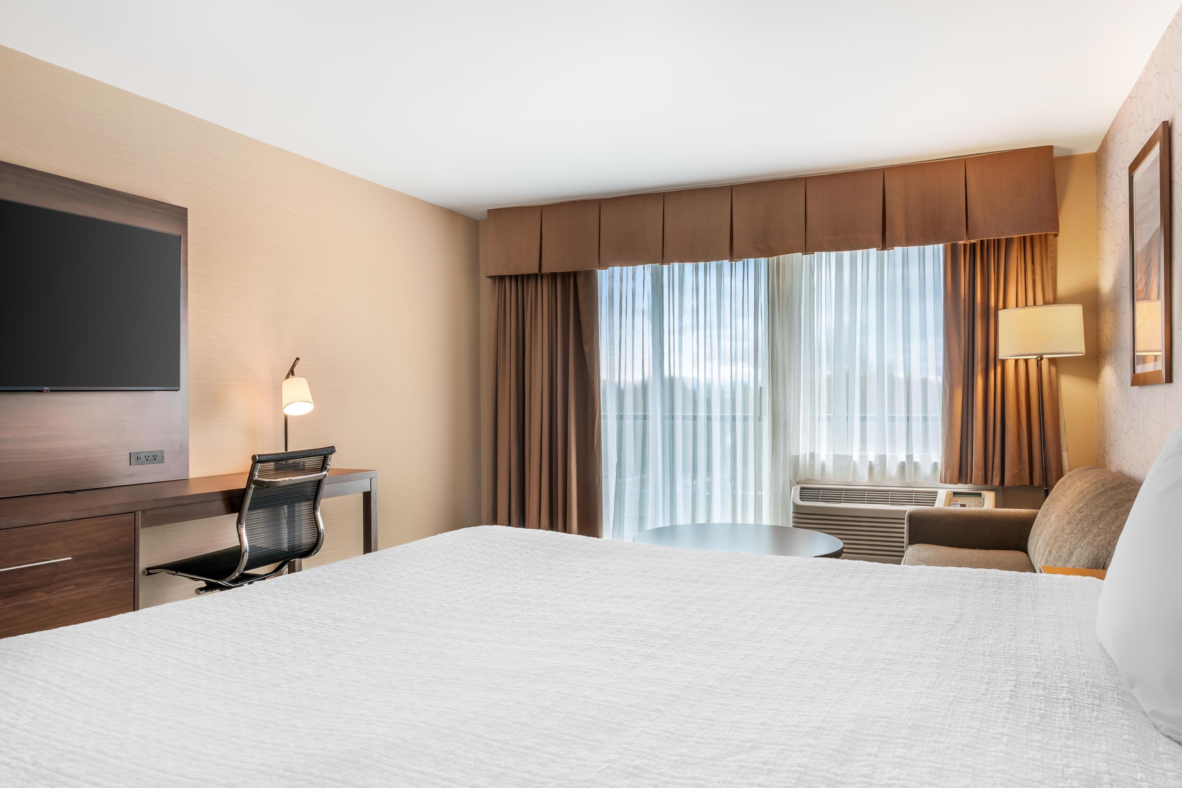 Habitación de hotel con cama, escritorio, silla, televisión y ventanas grandes con cortinas.