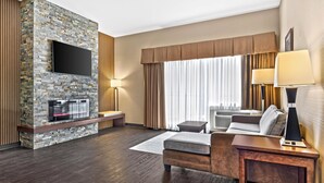 Una habitación de hotel moderna con chimenea de piedra, televisor de pantalla plana, sofá, mesa de centro y un amplio ventanal con cortinas.