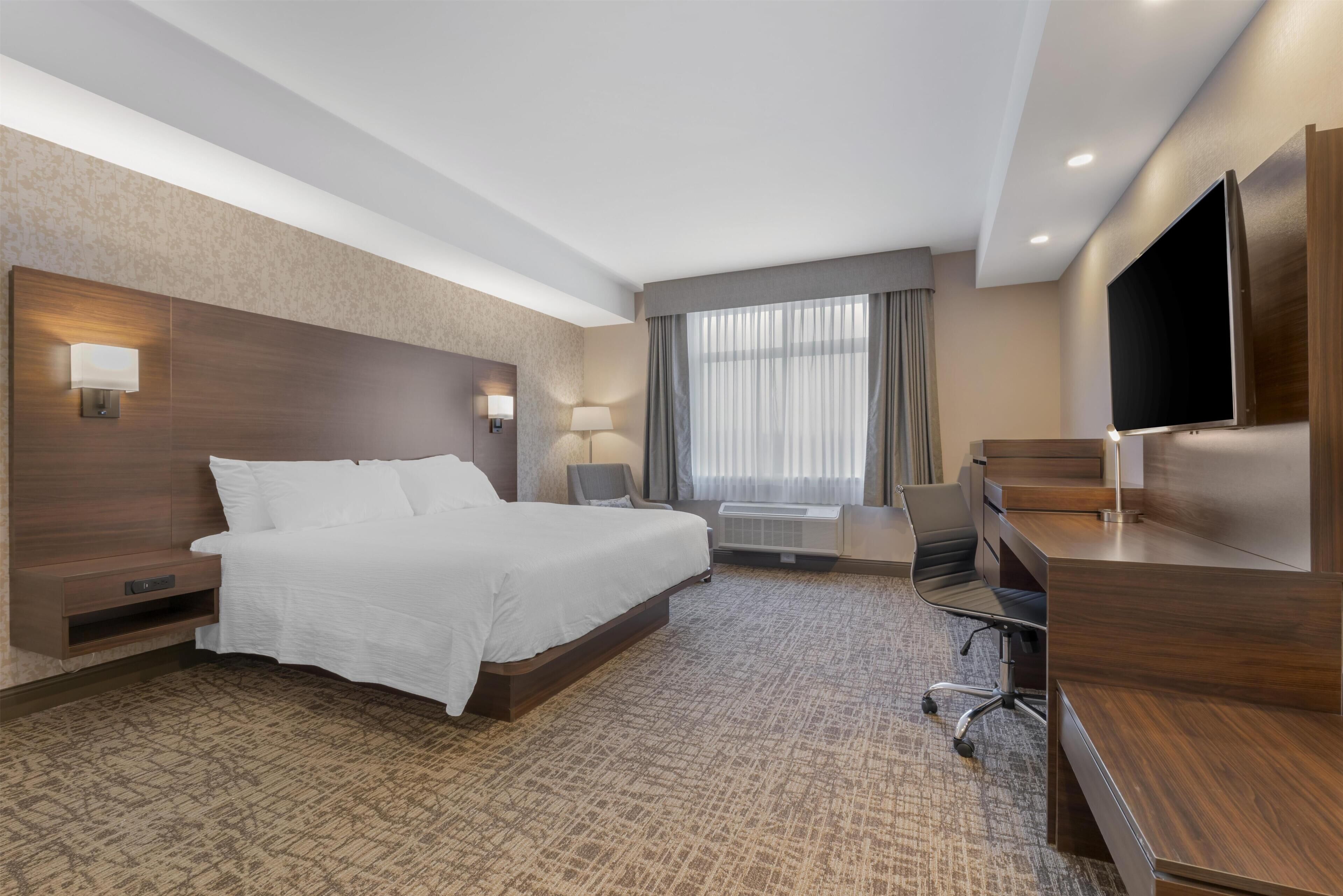 Ein Hotelzimmer mit einem großen Bett, einem Schreibtisch, einem Sessel, einem Fernseher und einem Fenster mit Vorhängen.