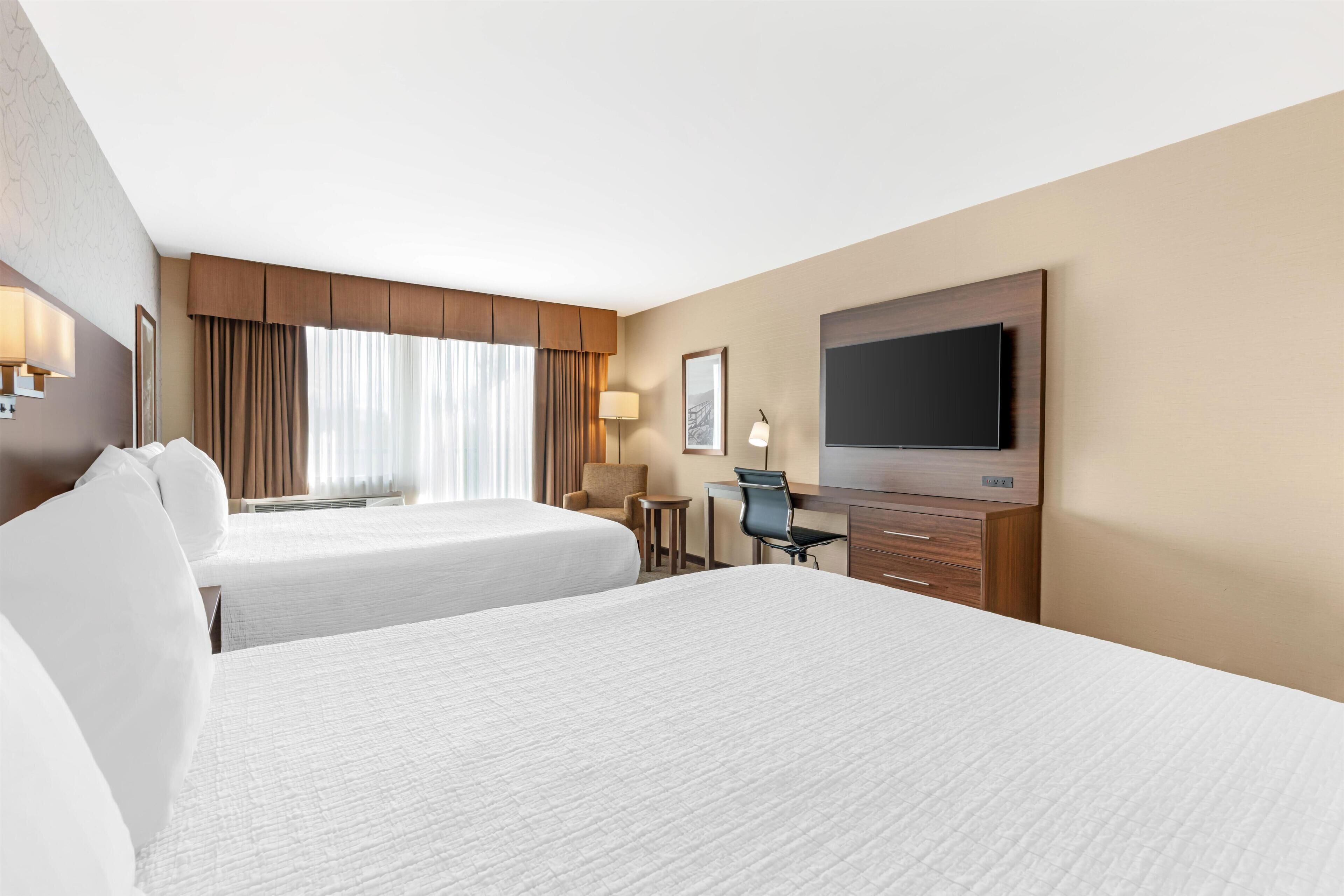 A hotel room with two beds, a desk, a chair, a TV, and a lamp.