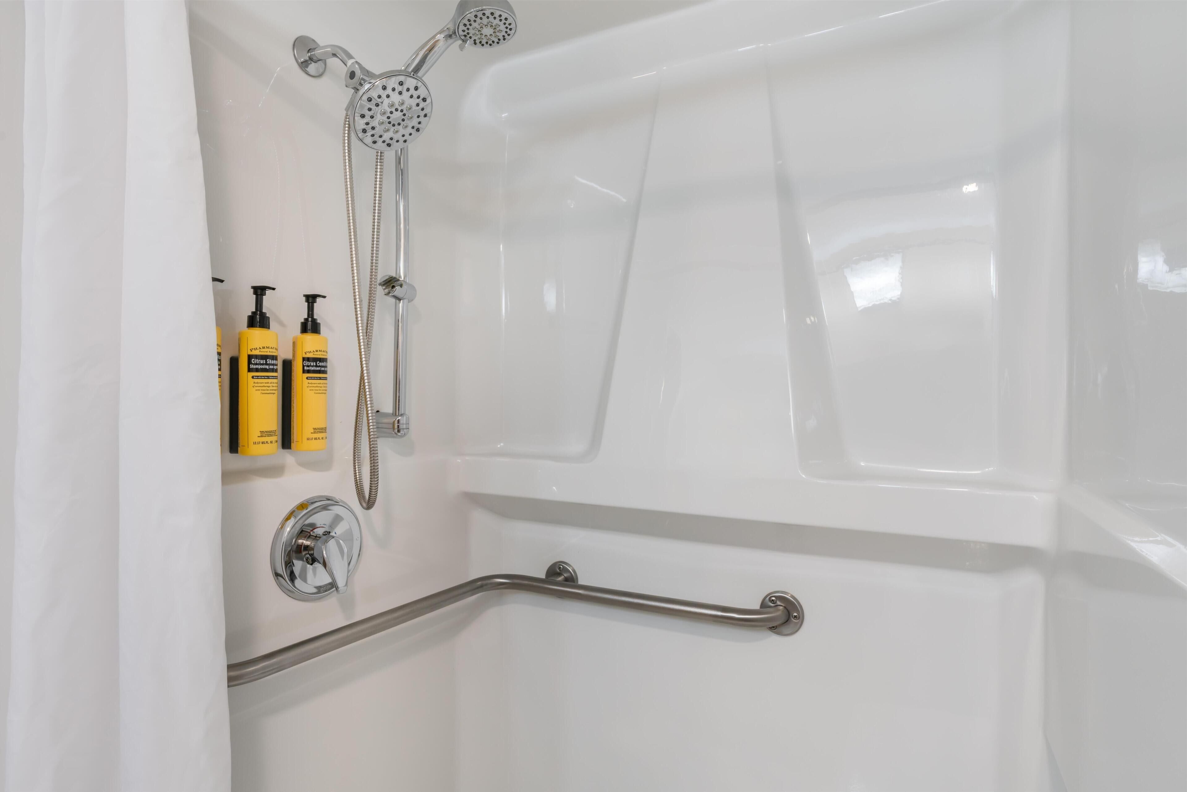 Une baignoire équipée d’un pommeau de douche à main, d’une barre d’appui et de deux flacons de shampoing jaunes.