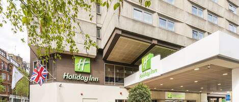 Un hotel Holiday Inn con fachada moderna, zona de estar exterior con flores coloridas y una bandera.