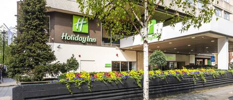 A Holiday Inn hotel with a modern facade, outdoor seating area with colorful flowers, and a tree in front.