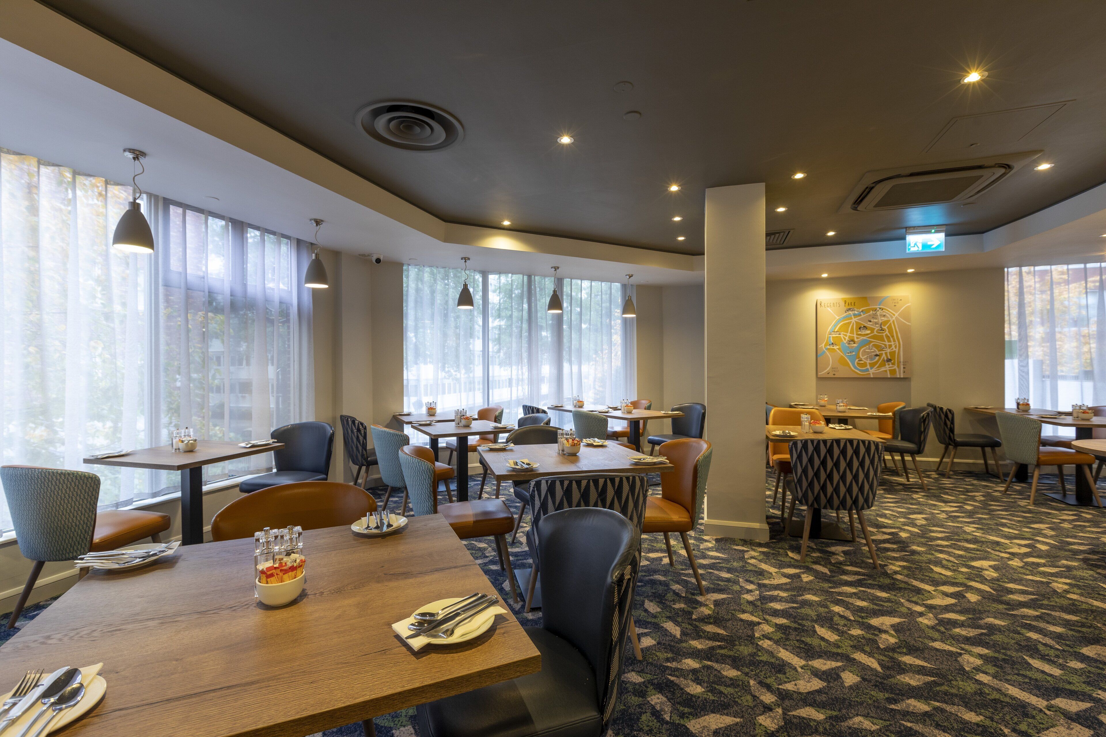 A hotel dining area with multiple tables and chairs, large windows with curtains, and a patterned carpet.