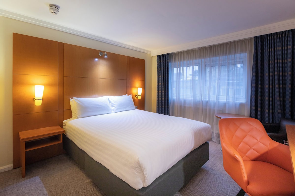 A hotel room with a large bed, bedside lamps, a small table, a chair, and a window with curtains.