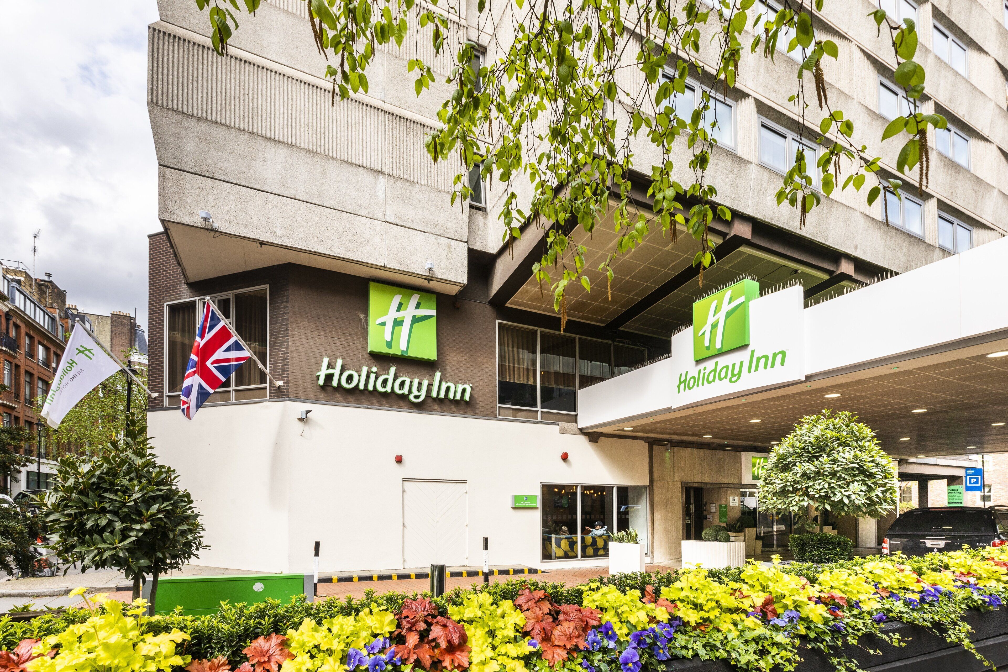 A Holiday Inn hotel with a modern facade, large windows, and a well-maintained entrance area with colorful flower beds.