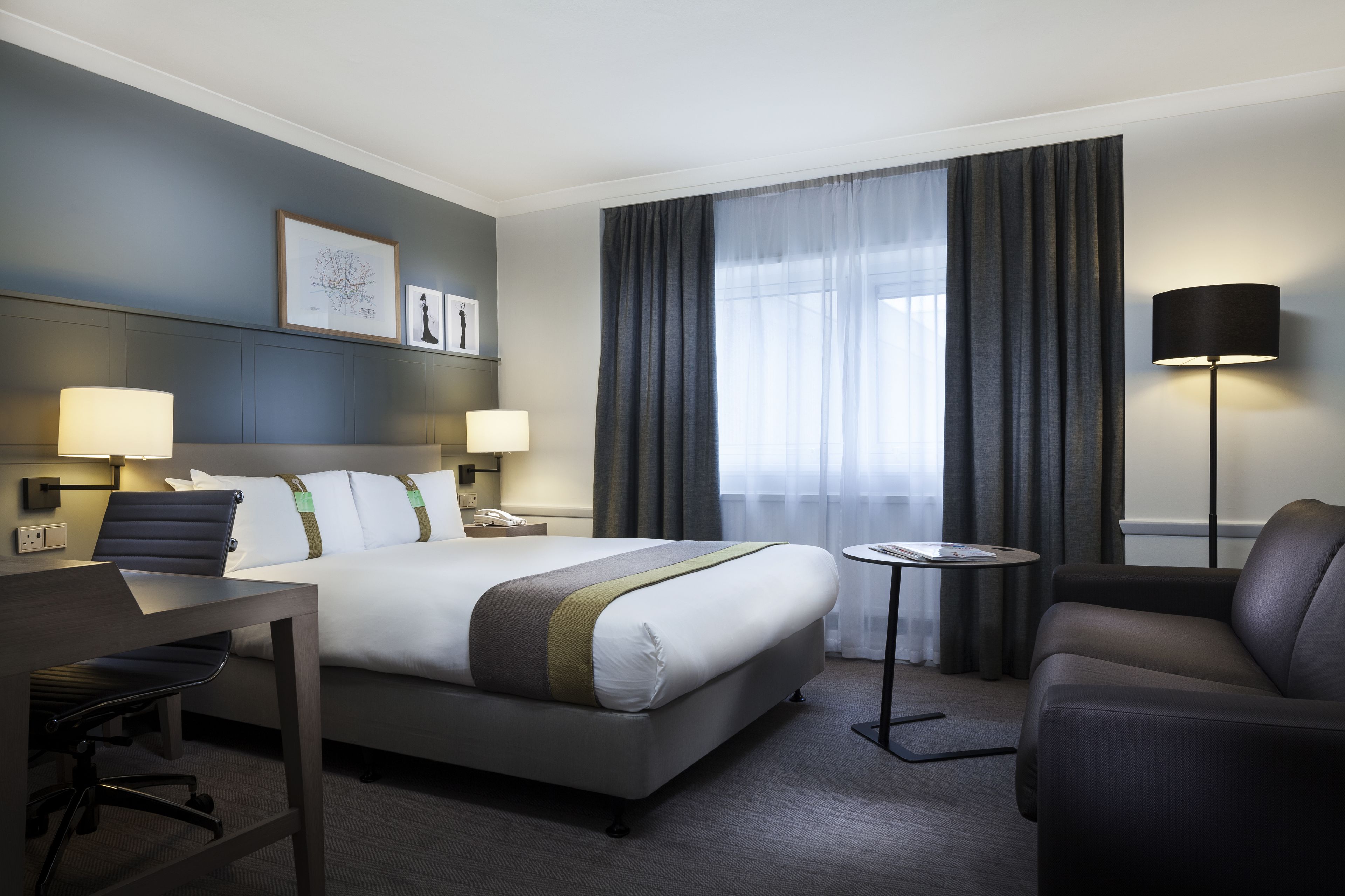 A hotel room with a bed, desk, chair, lamp, and artwork on the wall.