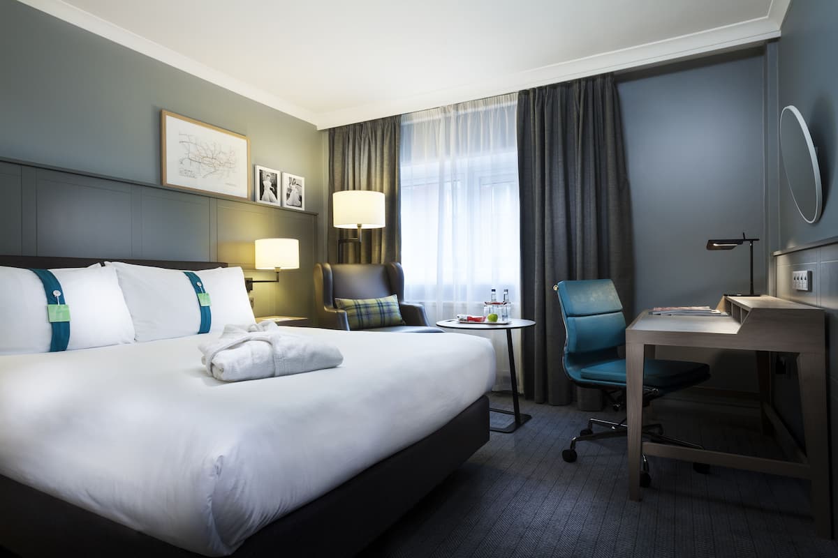A hotel room with a large bed, a desk, a chair, a lamp, and a window with curtains.