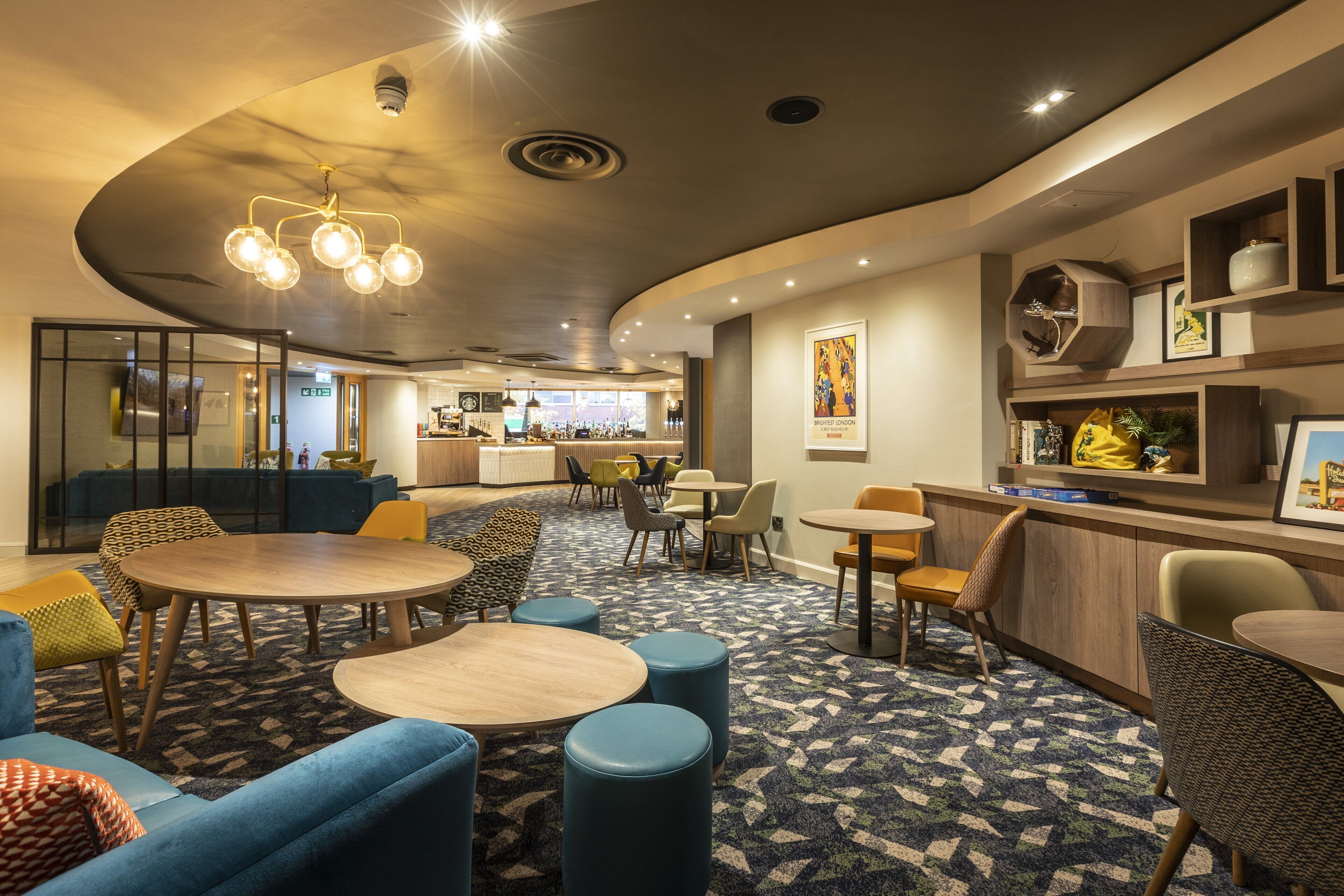 A modern hotel lobby with round tables, comfortable seating, a bar area, and a patterned carpet.