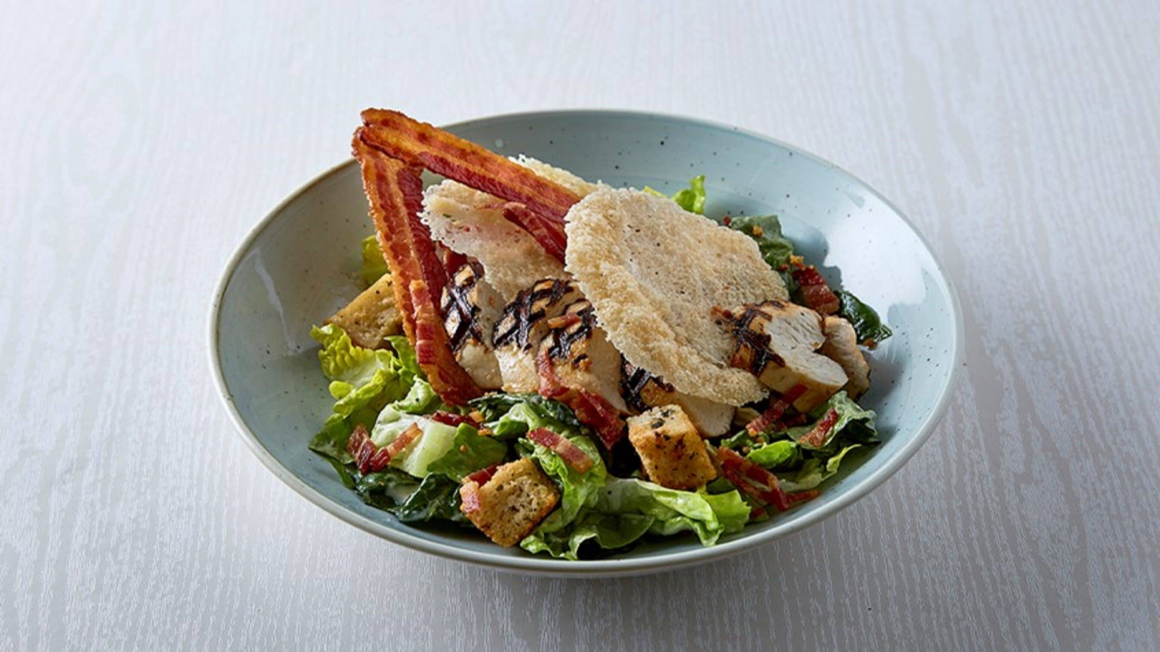 A bowl of salad with grilled chicken, bacon, croutons, and lettuce on a white surface.