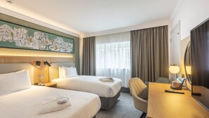 Une chambre d’hôtel avec deux lits, un bureau, une chaise, une lampe, un tableau au mur et une fenêtre avec des rideaux.