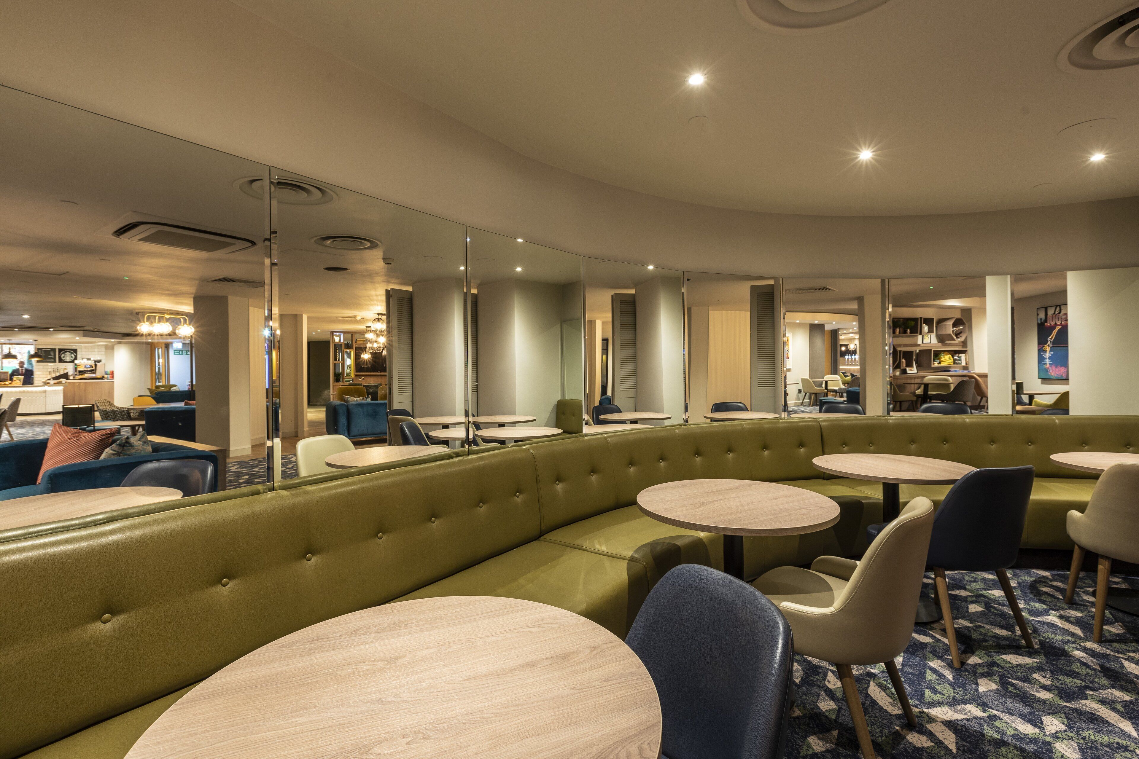 A modern hotel lobby with a green upholstered seating area, round tables, and a large mirror reflecting the space.