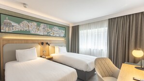 Une chambre d’hôtel avec deux lits, un canapé, un bureau avec une lampe, un tableau représentant un paysage urbain et une fenêtre avec des rideaux.