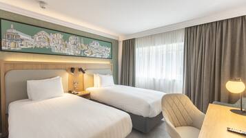 Een hotelkamer met twee bedden, een sofa, een bureau met lamp en een schilderij van een stadsgezicht, en een raam met gordijnen.