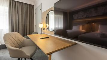 A hotel room with a wooden desk, a cushioned armchair, a large flat-screen TV, a round mirror, and a lamp.